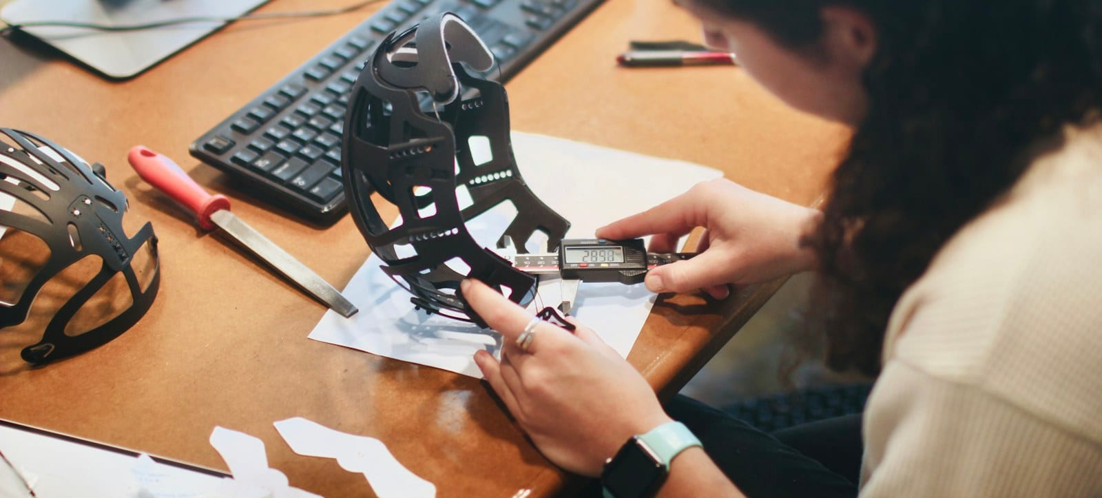 An inspector measures a black formed sheet metal part with digital calipers at a bench, the drawing beside it